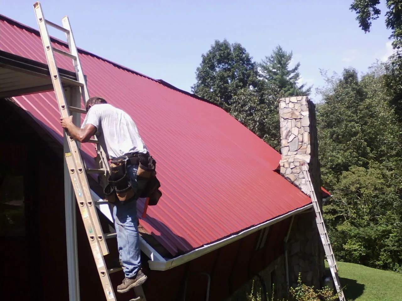Roofer on a ladder beside a red metal roof and stone chimney — Soffit & Fascia Repair by Roofing Brothers in Schnecksville